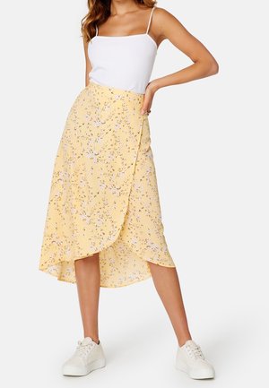 Kobieta w białym topie na cienkich ramiączkach, żółtej, kwiatowej spódnicy kopertowej i białych platformowych trampkach, stojąca z jedną ręką na biodrze.