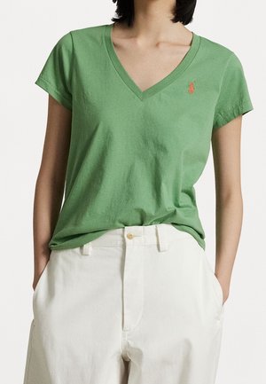 Kvinde iført en grøn V-hals T-shirt med korte ærmer og hvide bukser, hænderne i lommerne, vist fra skuldre til hofter.