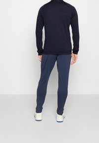 Giacca atletica blu navy con maniche lunghe e colletto alto, abbinata a pantaloni a gamba affusolata coordinati. Entrambi realizzati in un tessuto liscio e elasticizzato.