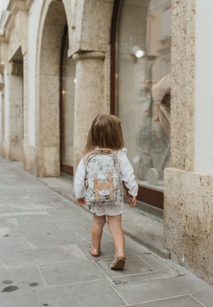 Zaino con motivo floreale e dettagli beige, forma arrotondata, materiale in tessuto, indossato da un bambino con abiti bianchi che cammina su un sentiero di pietra.