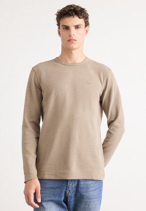 Beige long-sleeve T-shirt with a ribbed texture and round neckline, featuring a subtle logo on the left chest. Paired with blue jeans.
