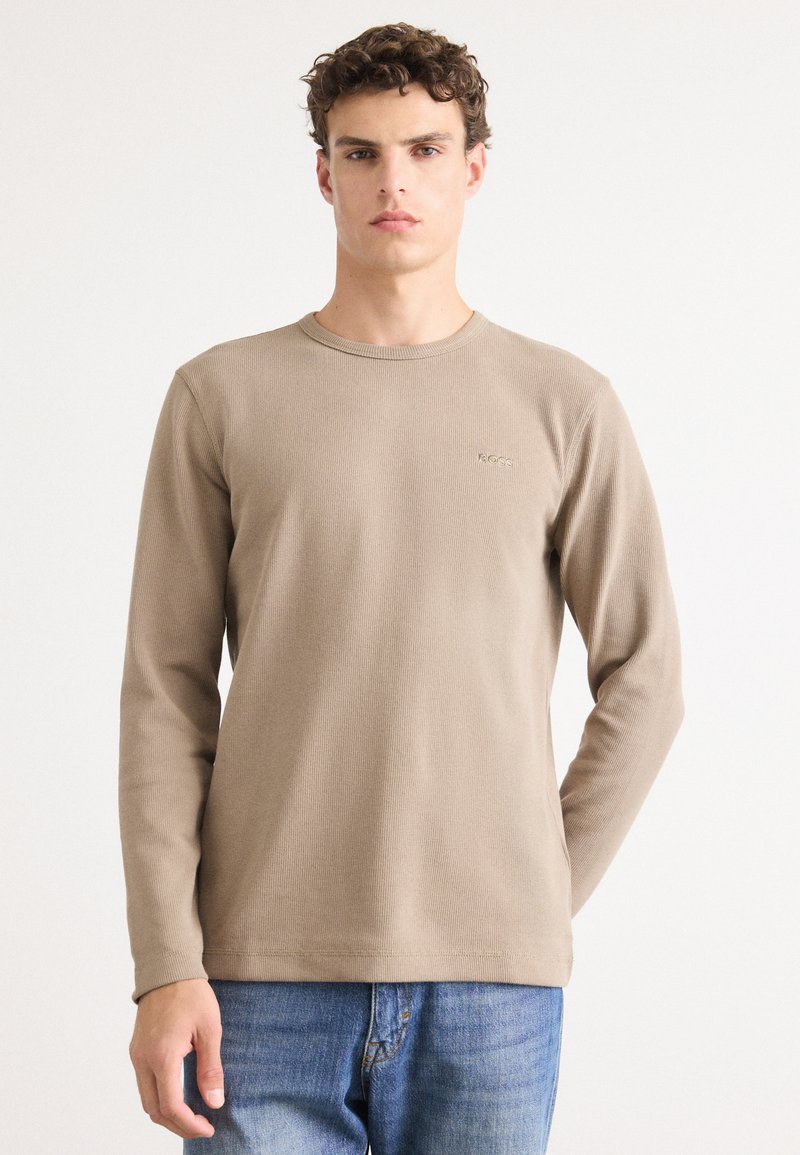 Beige lange mouwen T-shirt met een ribbelstructuur en ronde halslijn, voorzien van een subtiel logo op de linkerborst. Gekocht met blauwe jeans.
