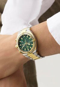 Kronografklocka med grön urtavla, guldindex, band av rostfritt stål i silver och guldton, samt strukturerade detaljer på bezeln.