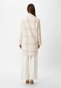 Lungo cappotto color crema con motivo a quadri rosa chiaro, realizzato in tessuto morbido, con colletto e maniche lunghe. Indossato sopra ampi pantaloni bianchi.