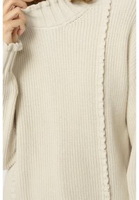 Cremefarbener Strickpullover mit gerafftem hohen Kragen und Rüschendetails entlang der Vorderseite und der Bündchen, eine Hand berührt sanft den Kragen.