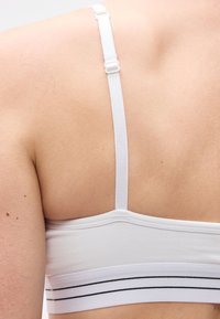 Κοντινή λήψη του επάνω μέρους της πλάτης ενός ατόμου που φοράει λευκό αθλητικό σουτιέν με ρυθμιζόμενες τιράντες και μαύρες οριζόντιες ρίγες στη βάση.