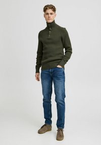 Rippene olivgrüne Pullover mit hohem Kragen, drei-Knopf-Leiste, kombiniert mit blauen Jeans und grauen Wildlederschuhen.
