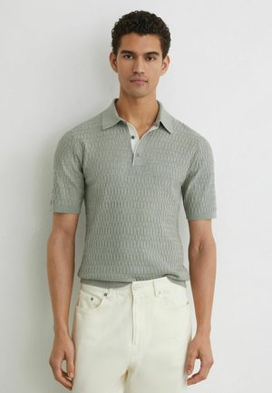 Mees, kes kannab lühikeste varrukatega tekstuuriga halli polo särki ja helevalgeid pükse, seisab ühetaolise heleda seina taustal, näoga ettepoole.