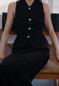 Veste sans manches noire avec fermetures à boutons dorés, dotée d'un col et d'une coupe ajustée. Texture lisse, associée à un bas long noir.