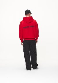 Röd hoodie med texten "CHAMP MODE" i svart på ryggen, kombinerad med svarta cargobyxor och vita sneakers. Enkelt design, avslappnad passform.