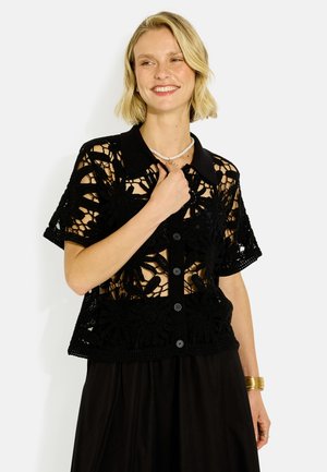 Mujer sonriente con cabello rubio corto viste un top negro de crochet de manga corta abotonado y falda negra con un collar blanco y una pulsera dorada.