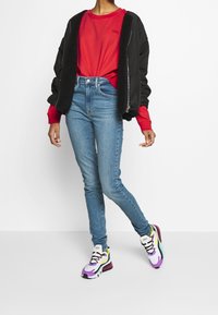 Czarna bomberka z kożuchem, czerwona bluza, niebieskie obcisłe dżinsy oraz wielokolorowe sneakersy z czarnymi akcentami i sznurowanym fasonem.