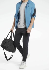 Felpa zipata azzurro chiaro con un pannello nero, t-shirt grigia, pantaloni neri a sigaretta, e una borsa da palestra nera, con scarpe da atletica bianche.