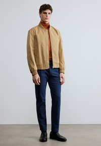 Beige bomberjacka med elastiska muddar, kombinerad med en orange polotröja, marinblå byxor och svarta läderskor. Slät textur, minimalistisk design.