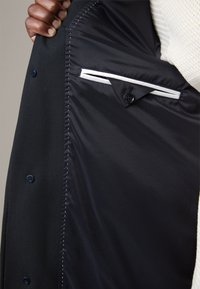 Binnenkant van een marineblauwe blazer met een gladde voering, een zakkie met witte bies en blauwe knopen. Gestructureerde stof aan de buitenkant.