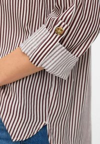 Gros plan sur une manche de chemise à rayures verticales marron et blanche, remontée et maintenue avec un bouton marron, portée avec un jean en denim bleu.