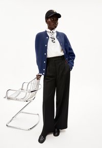 Marineblauwe cardigan met witte knopen over een wit shirt, zwarte broek en zwarte schoenen, gecombineerd met een zwarte pet met een gele opdruk.
