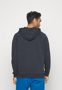 Mørk marineblå hettegenser med snor, raglanermer og ribbestrikkede mansjetter. Myk tekstur og kengurulomme foran. Båret med lyseblå bukser.