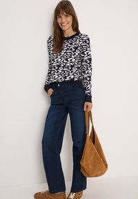 Pull naval avec imprimé animal blanc, jeans bleu foncé et sac à main en daim marron clair. Le modèle porte des baskets marron, posant devant un fond uni.