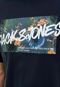 Lühikeste varrukatega tumesinine T-särk, millel on ristkülikukujuline graafika lillemustriga ja valge tekst "Jack & Jones" koos lisainfoga all.