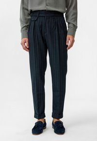 Marineblauwe gestreepte broek met plooien, voorzien van een ceintuur en omgeslagen broekspijpen, gemaakt van een gestructureerde stof. Ggedragen met donkerblauwe loafers.