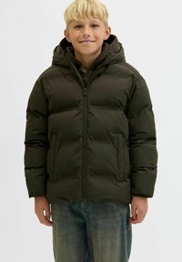 Lysblond dreng, der smiler, iført en mørkegrøn polstret jakke med hætte over en sort hoodie og blå jeans, stående mod en ensfarvet baggrund.