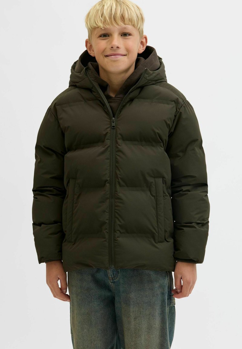 Lysblond dreng, der smiler, iført en mørkegrøn polstret jakke med hætte over en sort hoodie og blå jeans, stående mod en ensfarvet baggrund.
