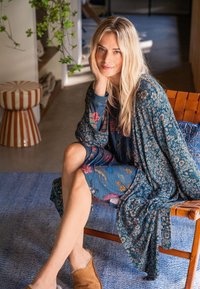 Blonde woman sitting on wooden chair, wearing blue floral dress and robe, resting chin on hand, with striped stool and green plant in background.