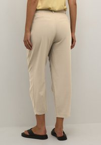 Pantalons crop de couleur crème avec une texture lisse, présentant un devant plat, deux poches arrière et un léger plissé. Portés avec des mules noires.