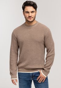 Brązowy sweter dzianinowy z teksturowanym wzorem, okrągłym dekoltem i białym wykończeniem przy mankietach oraz u dołu. Zestawiony z niebieskimi dżinsami.