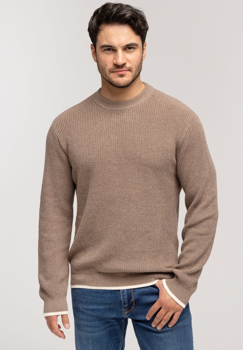 Brązowy sweter dzianinowy z teksturowanym wzorem, okrągłym dekoltem i białym wykończeniem przy mankietach oraz u dołu. Zestawiony z niebieskimi dżinsami.