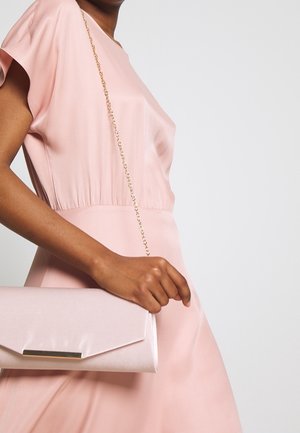 Donna in un abito di satin rosa pallido tiene in mano una pochette coordinata con una tracolla dorata su uno sfondo bianco semplice.