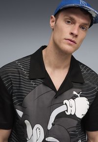 Kortærmet sort skjorte med grafisk design med striber og en tegneseriefigur. Sorte knapper og en åben krave fuldender looket.