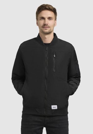 Black bomber jacket in smooth material with ribbed cuffs and hem. Features front zipper and chest pocket. Worn over a black shirt.