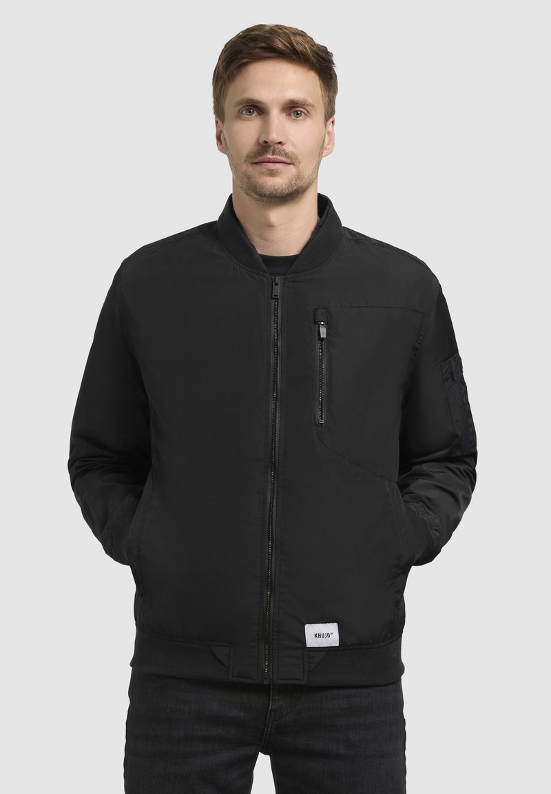 Schwarze Bomberjacke aus glattem Material mit gerippten Bündchen und Saum. Verfügt über einen Reißverschluss auf der Vorderseite und eine Brusttasche. Trägt man über einem schwarzen Hemd.