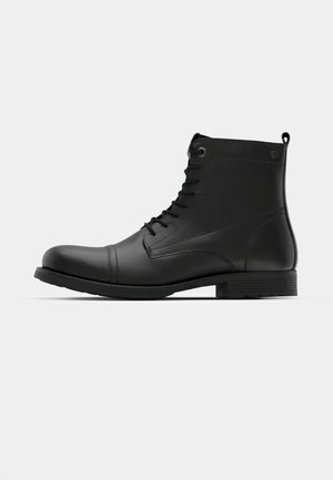 Botas de cuero negro hasta el tobillo con puntera redondeada, diseño con cordones y suela de goma plana. Presentan costuras mínimas y un lazo en el talón.
