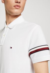 Vit polo skjorta med klassisk krage, försedd med en liten logotyp på bröstet och kontrasterande ränder i röd och marinblå på ärmarna.