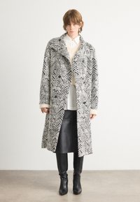 Abrigo largo de patrón de espiga en blanco y negro, con un frente de doble botonadura, cuello ancho y textura suave. Llevado sobre una blusa blanca.