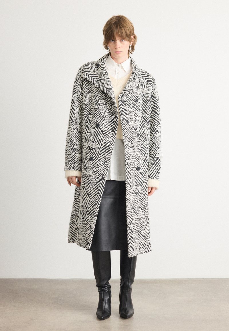 Cappotto lungo a motivo houndstooth in bianco e nero, con frontale doppiopetto, ampio colletto e texture morbida. Indossato sopra una maglietta bianca.