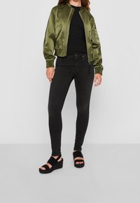 Chaqueta bomber de satén verde oliva con cierre de cremallera, camiseta negra ajustada, jeans negro ajustados y sandalias de cuña negras con dos tiras.