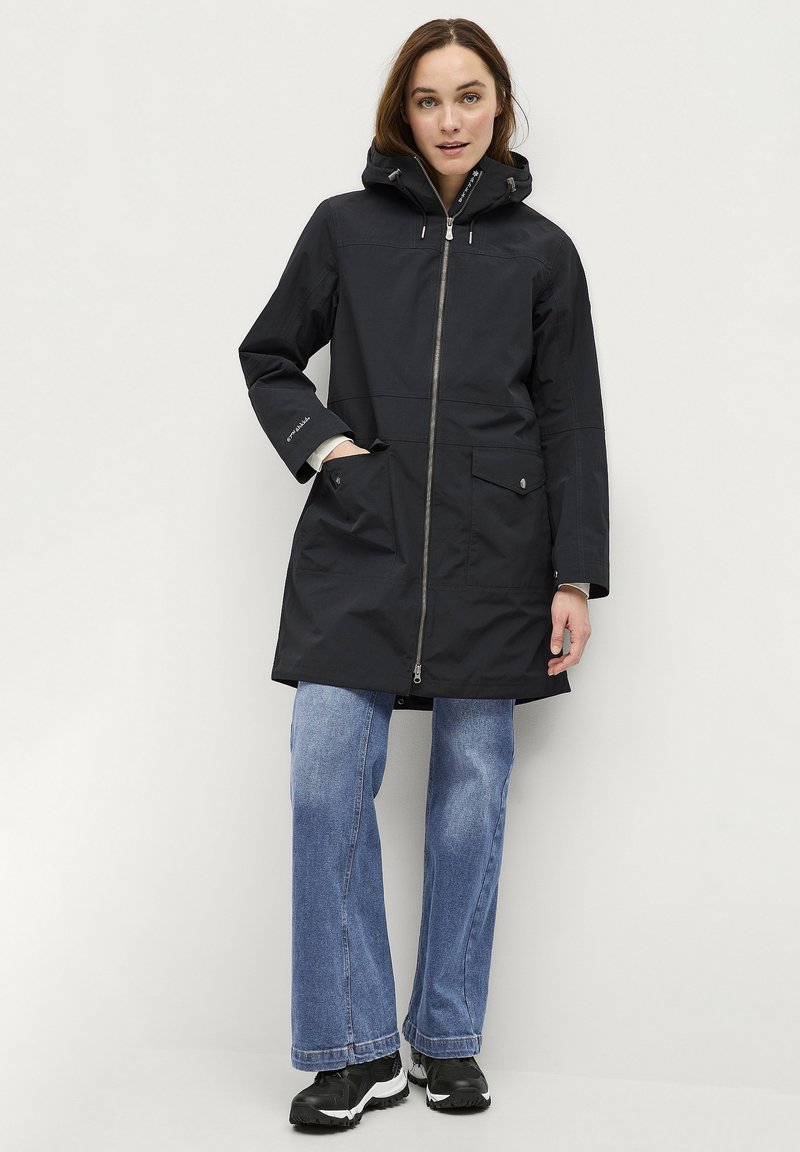 Parka noir imperméable avec fermeture éclair à l'avant, grandes poches et capuche. Présente une coupe décontractée et est associée à un jean bleu clair et des chaussures noires.