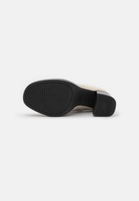 Zapato beige con un upper sintético texturizado, una suela de goma negra y un tacón cuadrado. La suela presenta un patrón de relieve en zigzag.