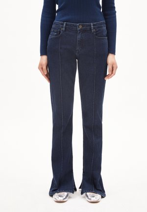Frau trägt dunkelblaue, ausgestellte Jeans mit vorderen Nähten, kombiniert mit silbernen Schuhen mit quadratischer Spitze und einem marineblauen, gerippten Pullover.