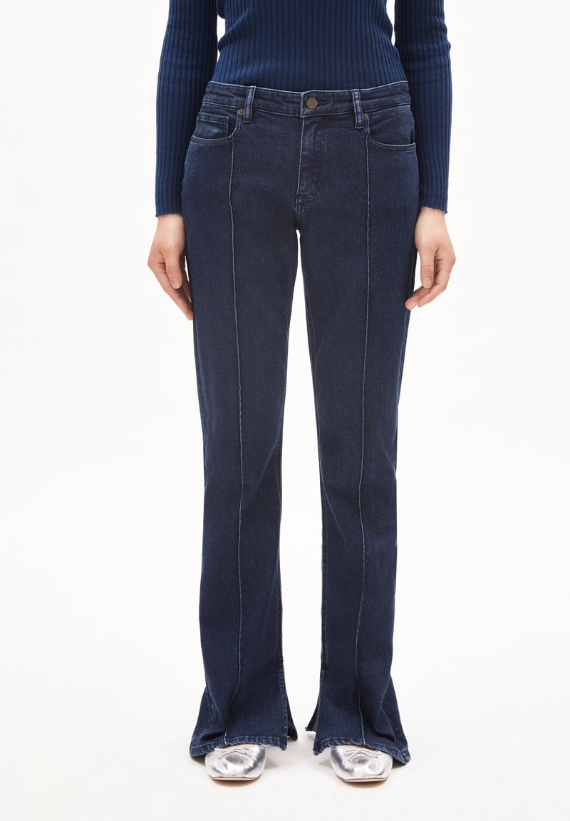 Femme portant un jean flare bleu foncé avec des coutures devant, assorti à des chaussures argentées à bout carré et un pull côtelé bleu marine.
