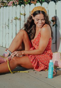 Różowe wrotki z żółtymi sznurowadłami, czerwona sukienka w kwiatowy wzór, obok butelka turkusowego sunscreenu, a w tle biała płot. 