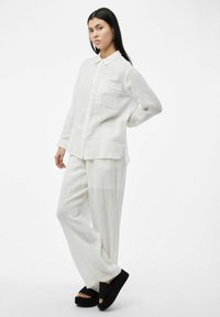 Camisa ligera de lino blanco con cuello y bolsillo, combinada con pantalones sueltos a juego. Se lleva con sandalias de plataforma negras.