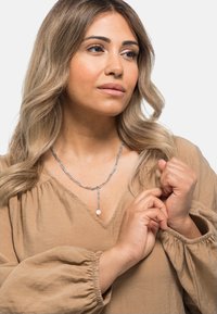 Silberne Kettenhalskette mit länglichen Gliedern und einem runden Anhänger. Das Modell trägt eine beige Bluse mit Puffärmeln. Weiche Textur.