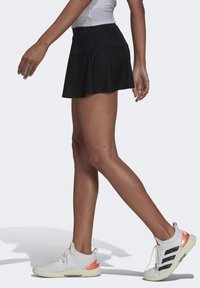 Una falda corta atlética negra con un diseño en capas, que se lleva con zapatos blancos con acentos en naranja y negro. Textura suave y cintura ajustada.