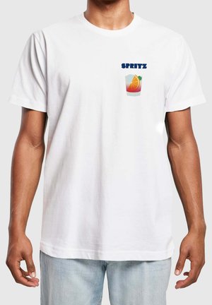 Hvid t-shirt med lille grafik af et glas med appelsinskive og ordet "SPRITZ" i blåt på venstre bryst, båret af en person i lyse jeans.
