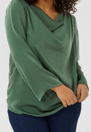 Femme portant un haut vert ample à manches longues avec un décolleté drapé, tenant le bas du haut, associé à un pantalon bleu foncé.